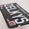 A black license plate with white lettering and pink numbers.
